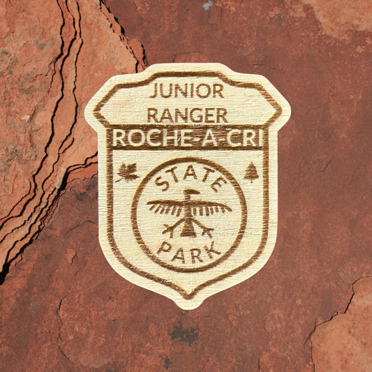 Wood Shaped badge with the text “Junior Ranger Roche-A-Cri State Park” and an petroglyph symbol.  Photo background: rock.