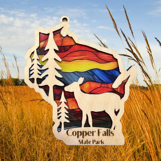Wood Wisconsin shaped ornament with trees, deer, and the text “Copper Falls State Park.”  Features a stained glass like background.  Photo background: tall grasses in fall.