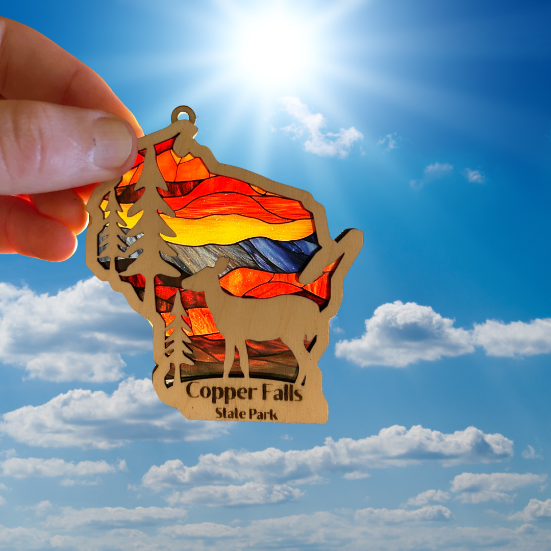 Wood Wisconsin shaped ornament with trees, deer, and the text “Copper Falls State Park.”  Features a stained glass like background.  Photo background: ornament held up to sun.