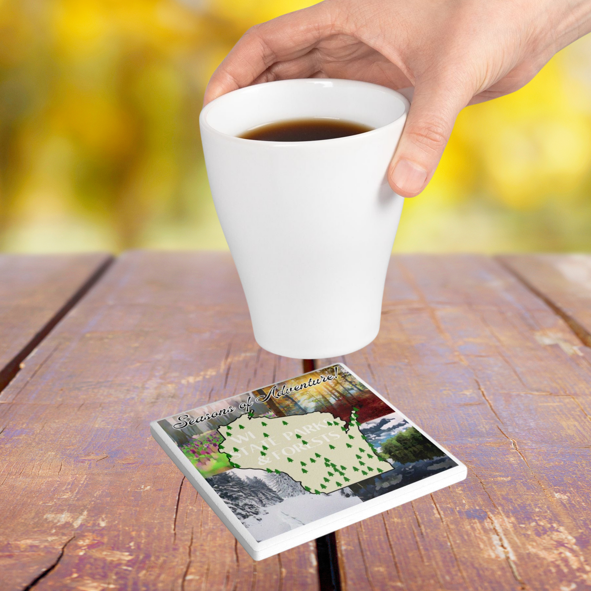 Ceramic coaster featuring a map of all the parks and forests of Wisconsin on a background depicting images of different seasons of winter, spring, summer, and fall.  Photo background: a hot beverage being set on a coaster that is on a rustic picnic table.