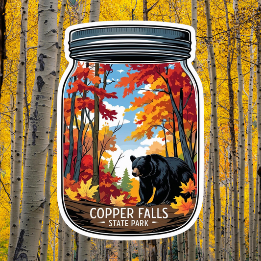 A mason jar shaped decal featuring a design of a bear walking through an autumn woods and the text Copper Falls State Park. Photo background, birch trees with bright yellow foliage.
