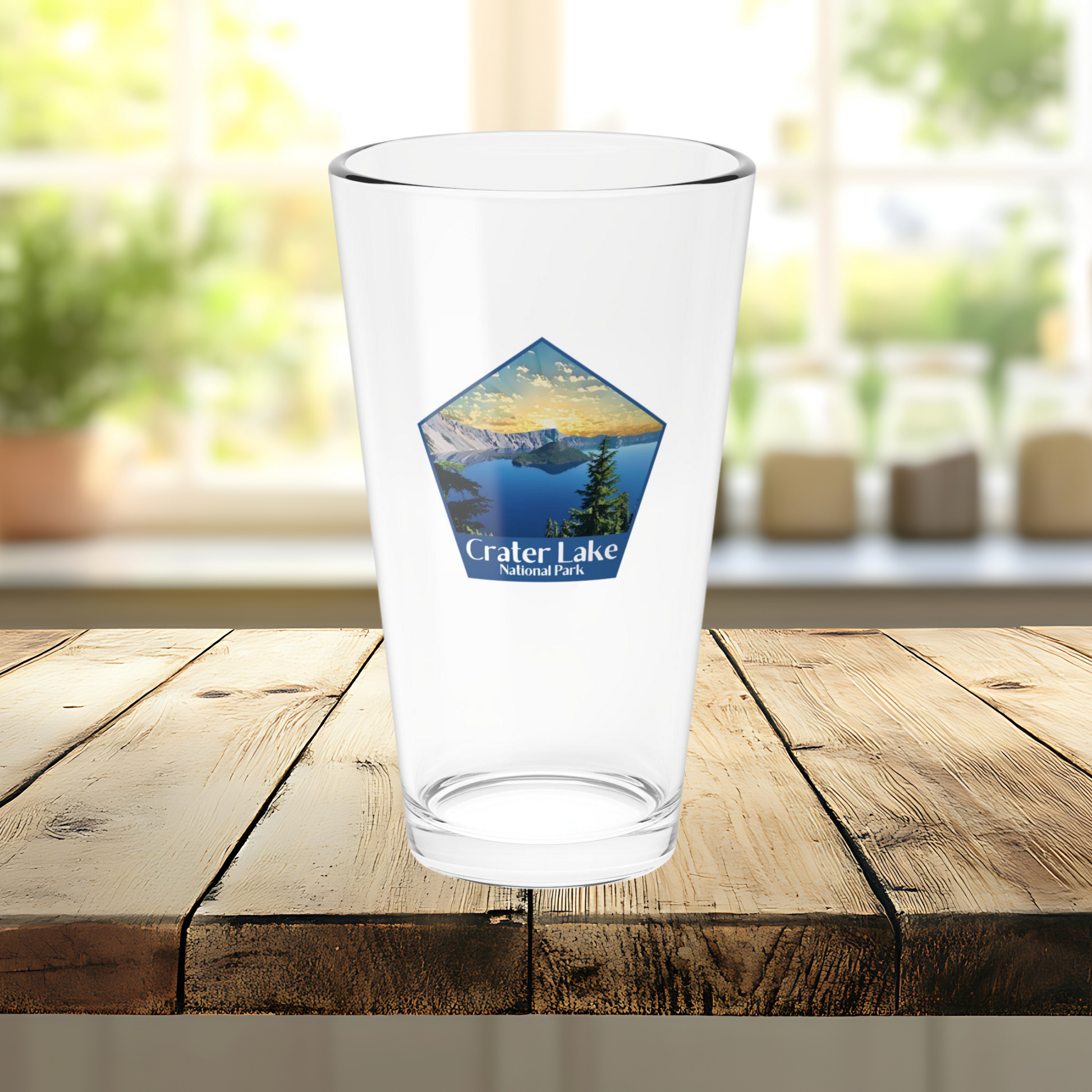 16oz pint glass sitting on a wood counter with plants on a window sill in the background.  Pint glass features a pentagon shaped design of a sunrise over Crater Lake and the Text Crater Lake National Park.