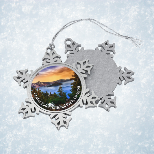 Pewter snowflake ornaments front and back against a snowflake background.  Ornament features a vivid sunrise over Crater Lake design.