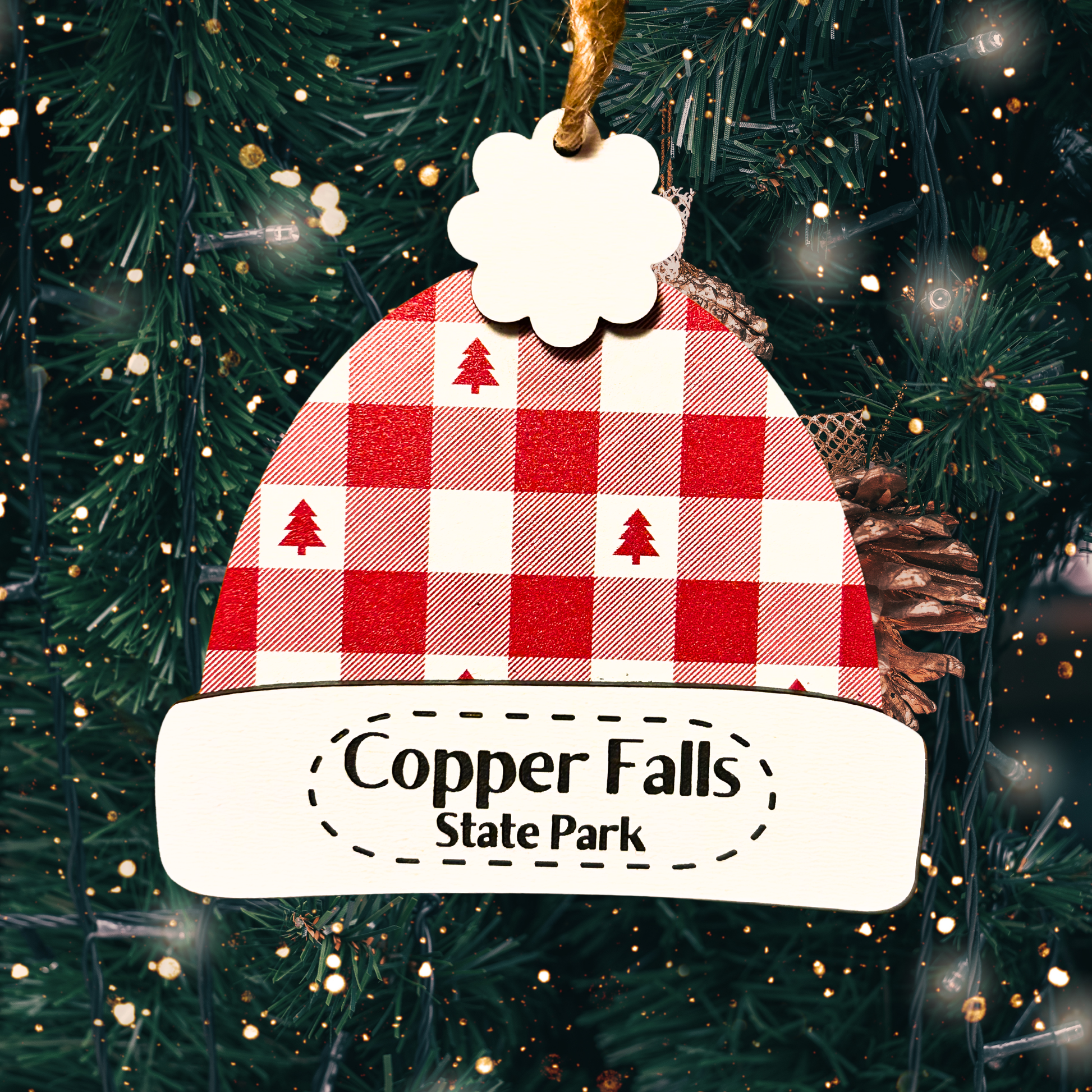 Red and white plaid  with red tree shape patterned beanie hat ornament with 'Copper Falls State Park' text engraved on the ornament.  Photo background of a close up view of a lighted Christmas tree.