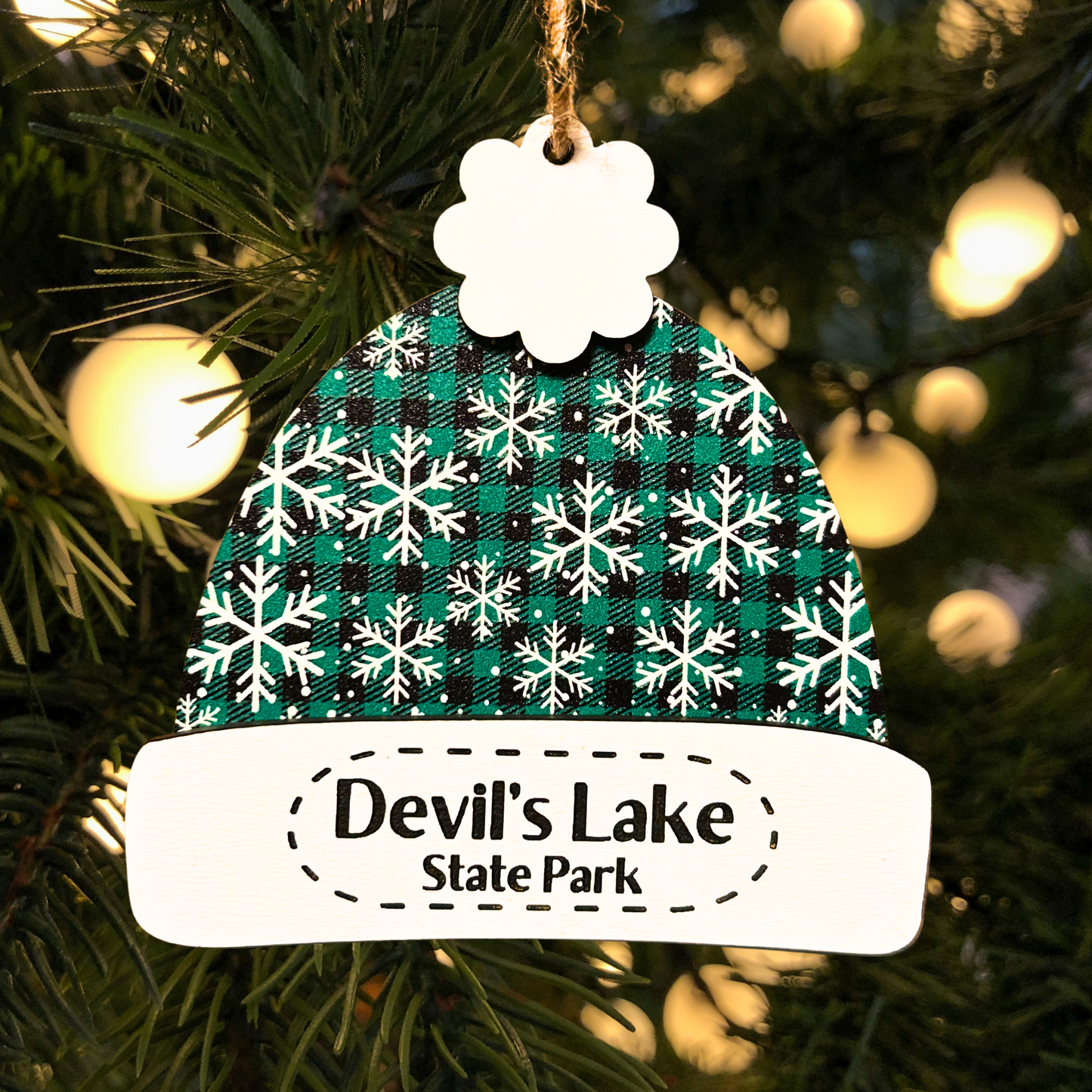 Decorative winter beanie hat ornament with a green and black plaid with snowflake pattern and 'Devil's Lake State Park' text engraved in the ornament.  Photo background: close up of a Christmas tree and lights.