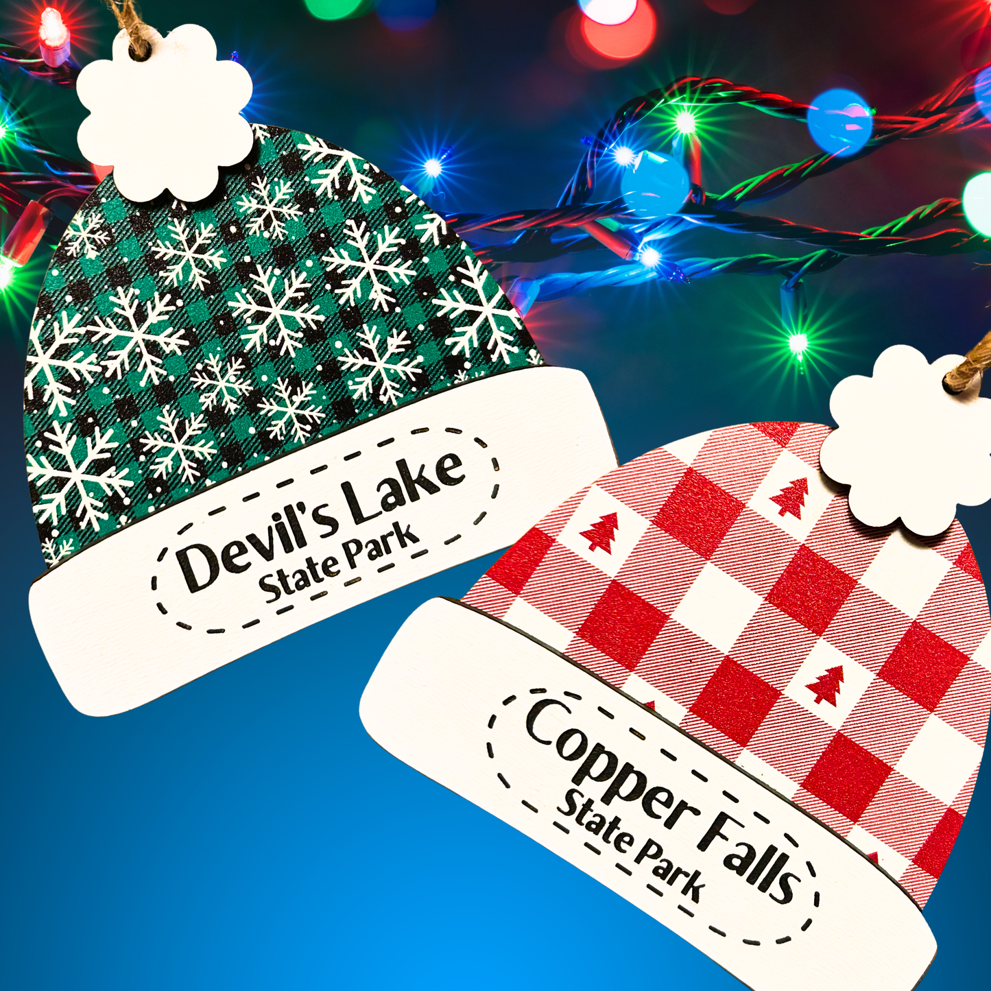 Two Christmas-themed winter beanie hat ornaments with 'Devil's Lake State Park' and 'Copper Falls State Park' text against a festive background. One ornament is a red and white plaid pattern with red Christmas trees, and the other ornament is a green and black plaid pattern with white snowflakes.