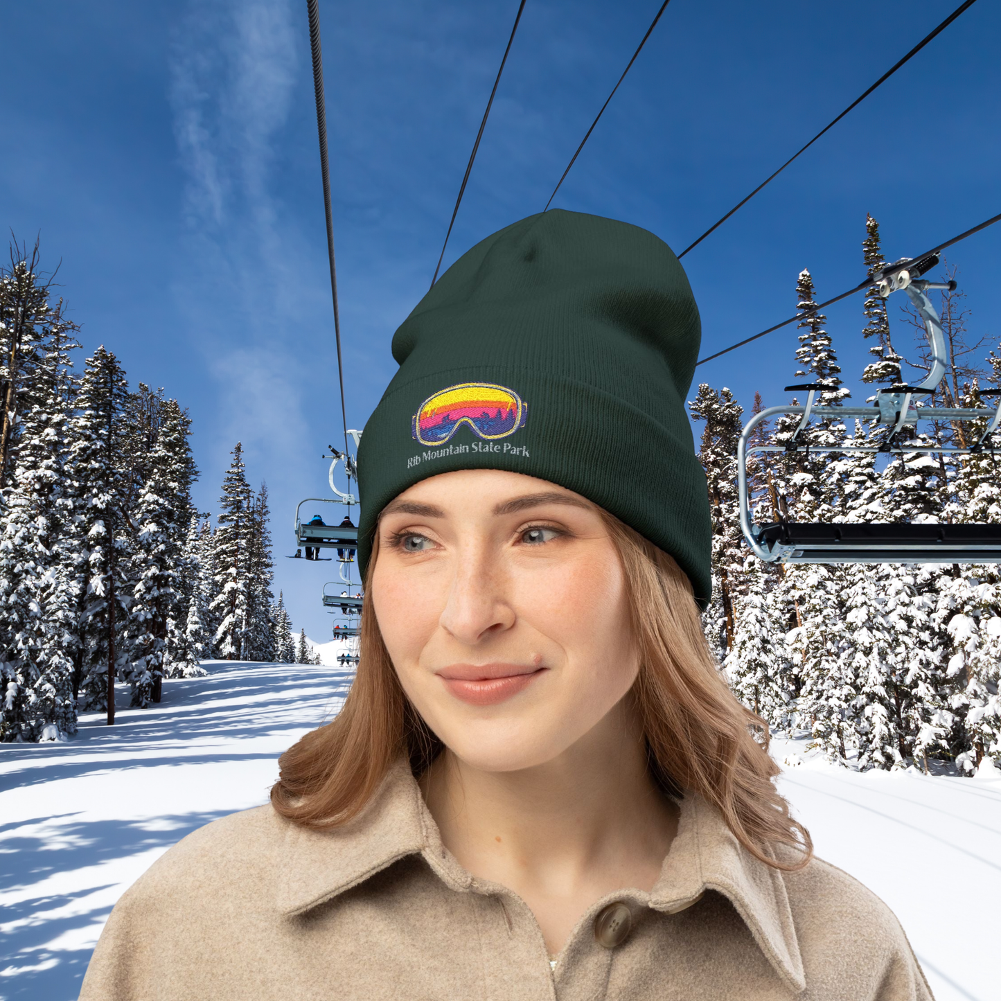 Woman wearing a dark green beanie with with colorful goggles design and Rib Mountain State Park text against a snowy ski lift background