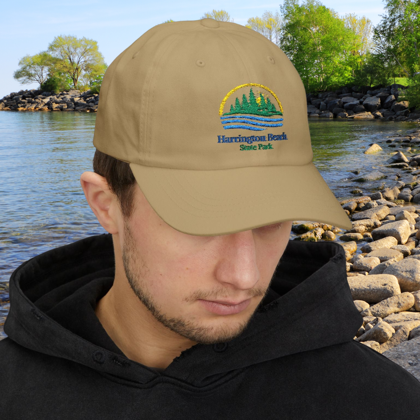 Man wearing a khaki cap with Harrington Beach State Park logo, standing by a body of water with rocks and trees.
