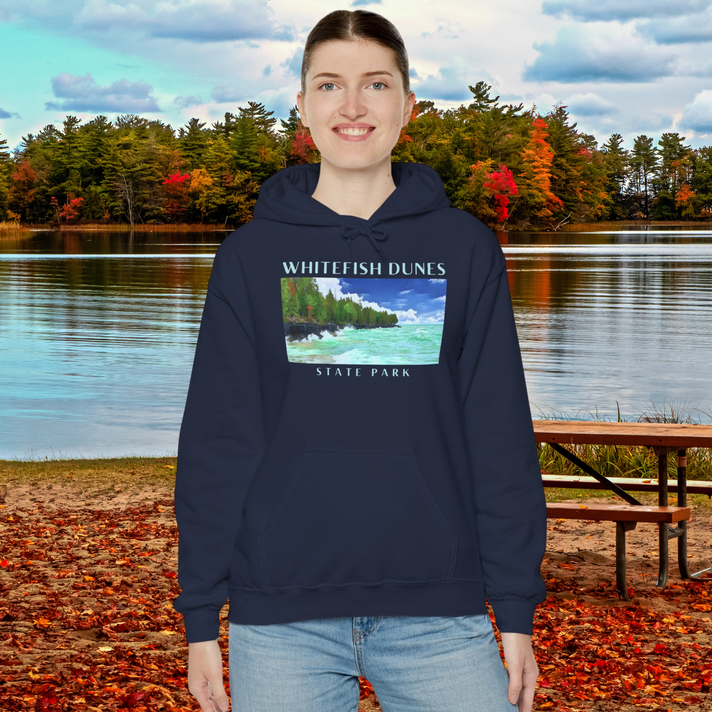 Person wearing a navy hoodie with Whitefish Dunes State Park design, standing in front of a lake with autumn foliage.