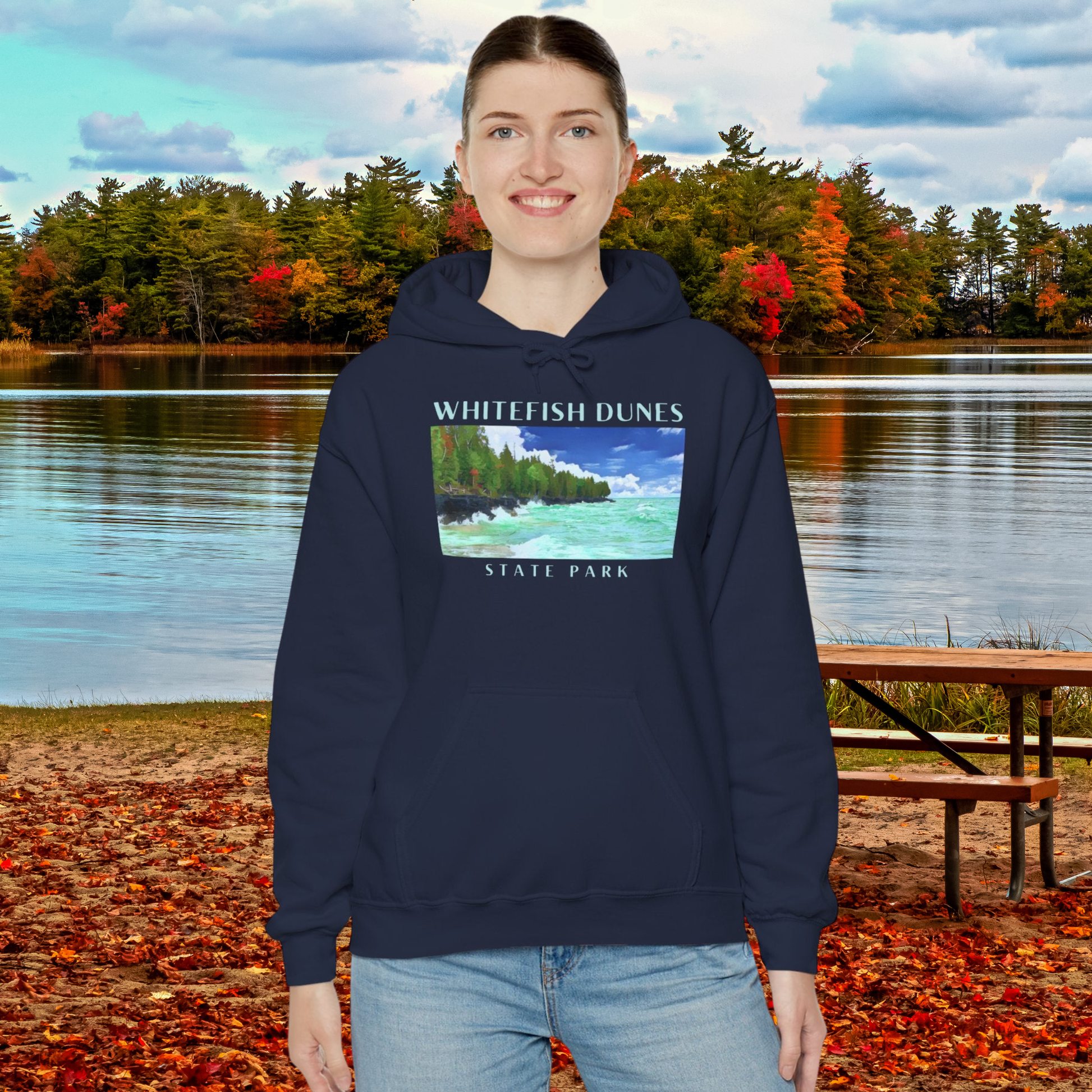 Person wearing a navy hoodie with Whitefish Dunes State Park design, standing in front of a lake with autumn foliage.