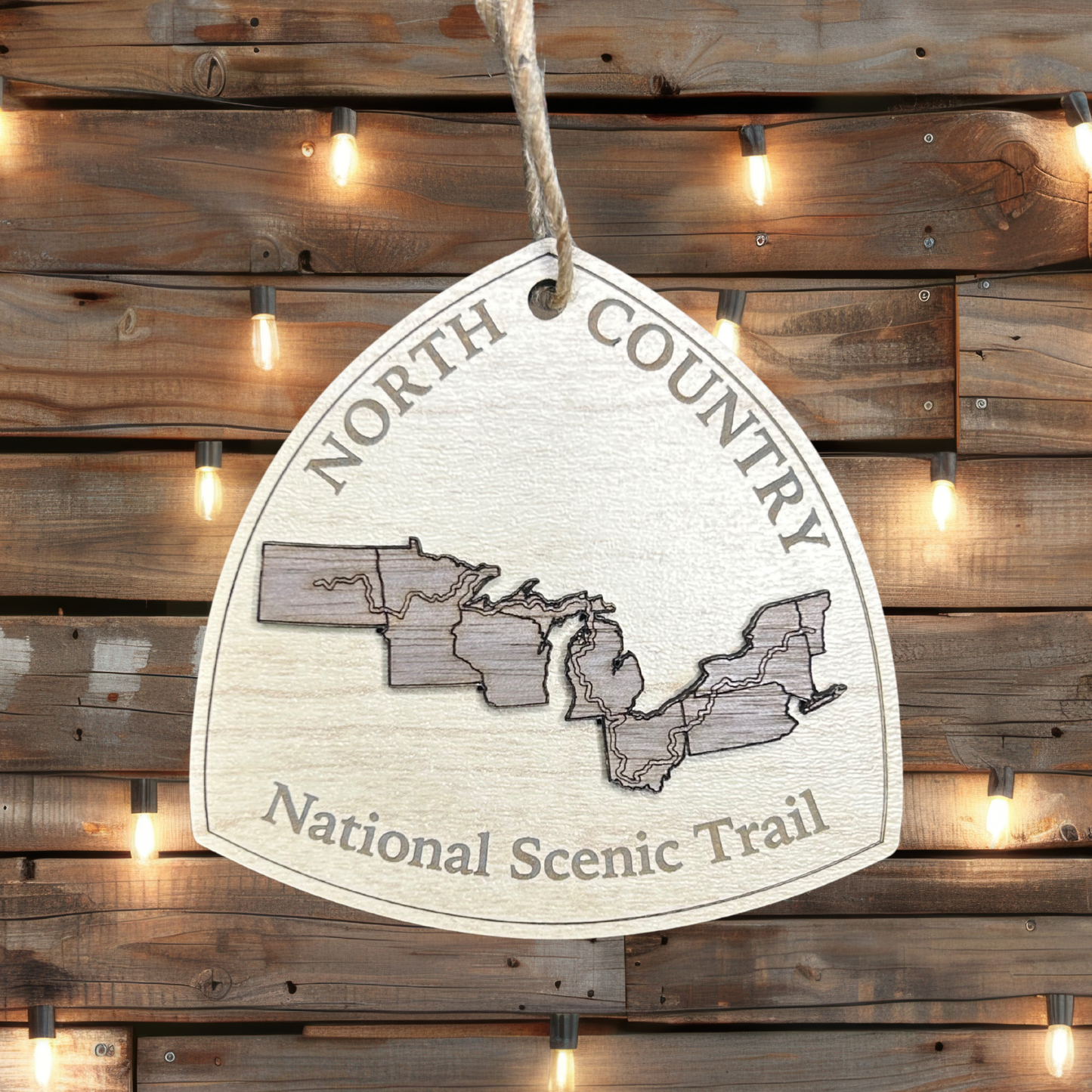Wood ornament in the shape of the national trail's icon, with a walnut North Dakota, Minnesota, Wisconsin, Michigan, Ohio, Pennsylvania, New York, and Vermont Shaped center piece depicting the North Country Trail Route.  Ornament contains the text "North Country National Scenic Trail".  Photo background: rustic wood wall with lights.