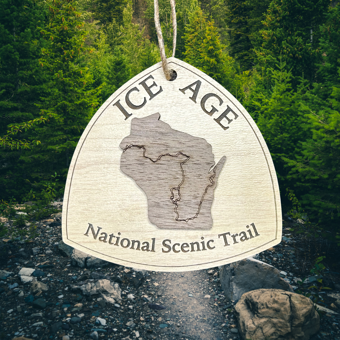 Wood ornament in the shape of the national trail's icon, with a walnut Wisconsin Shaped center piece depicting the Ice Age Trail Route.  Ornament contains the text "Ice Age National Scenic Trail".  Photo background: primative hiking trail through dense forest.