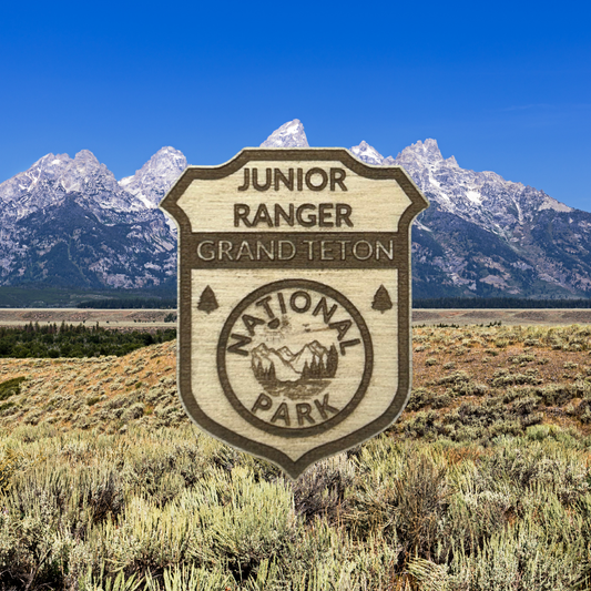 Wood badge featuring the text: “Junior Ranger Grand Tetons National Park” with a mountain and lake seal.  Photo background: snowcapped mountains in far off distance.