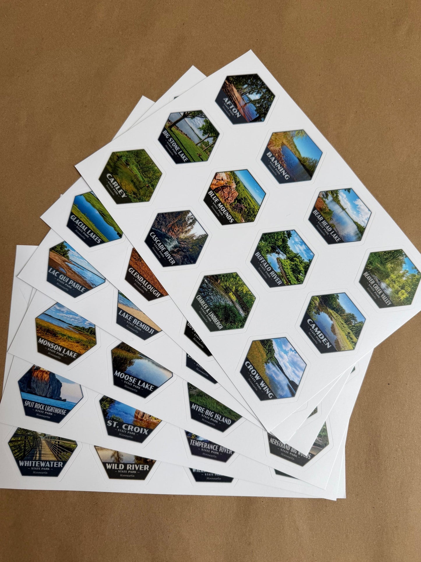 Several sheets of small hexagonal scenic stickers fanned out on a brown kraft paper background. Each sticker shows a different landscape photo of Minnesota’s State Parks.