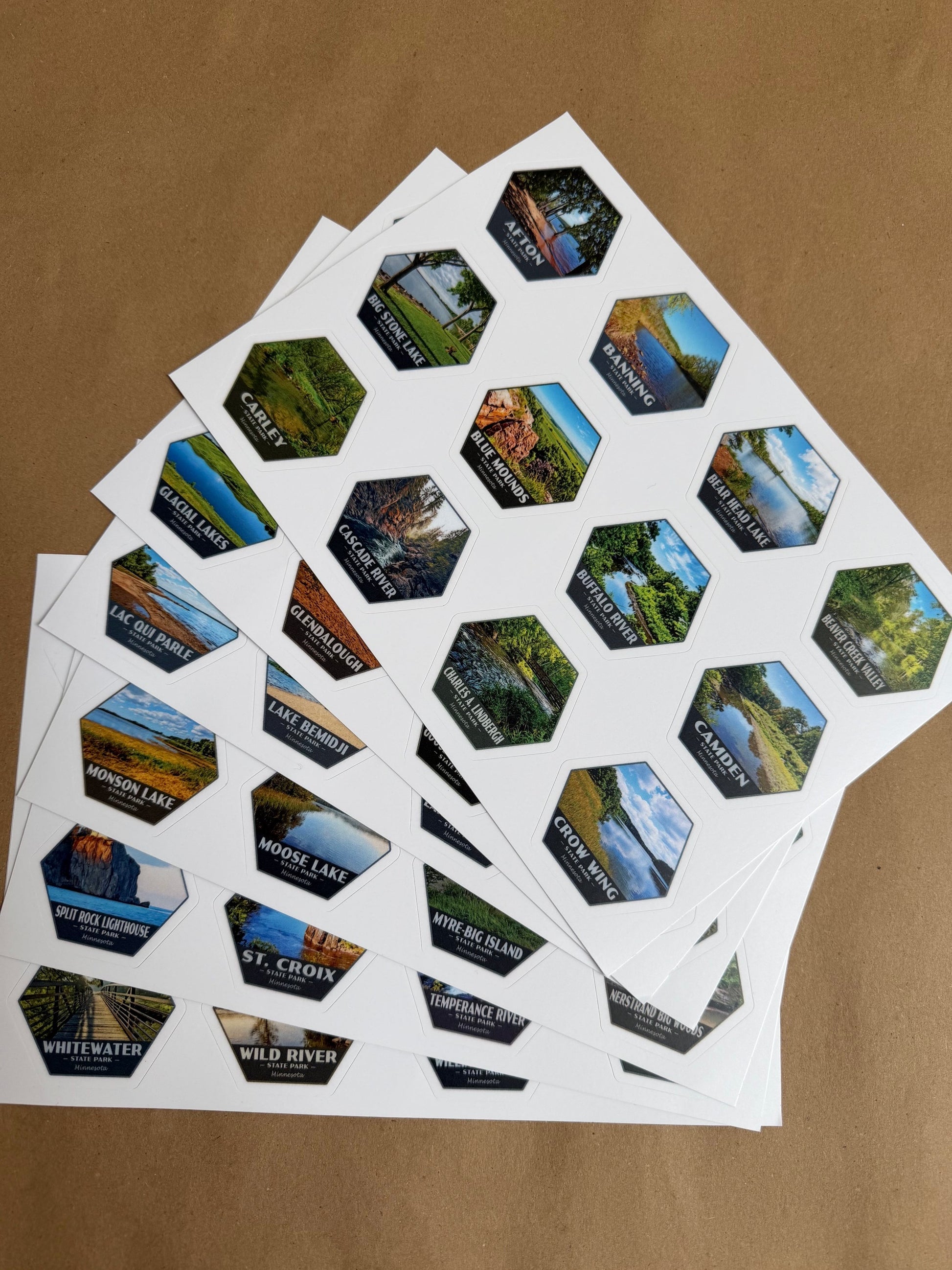 Several sheets of small hexagonal scenic stickers fanned out on a brown kraft paper background. Each sticker shows a different landscape photo of Minnesota’s State Parks.