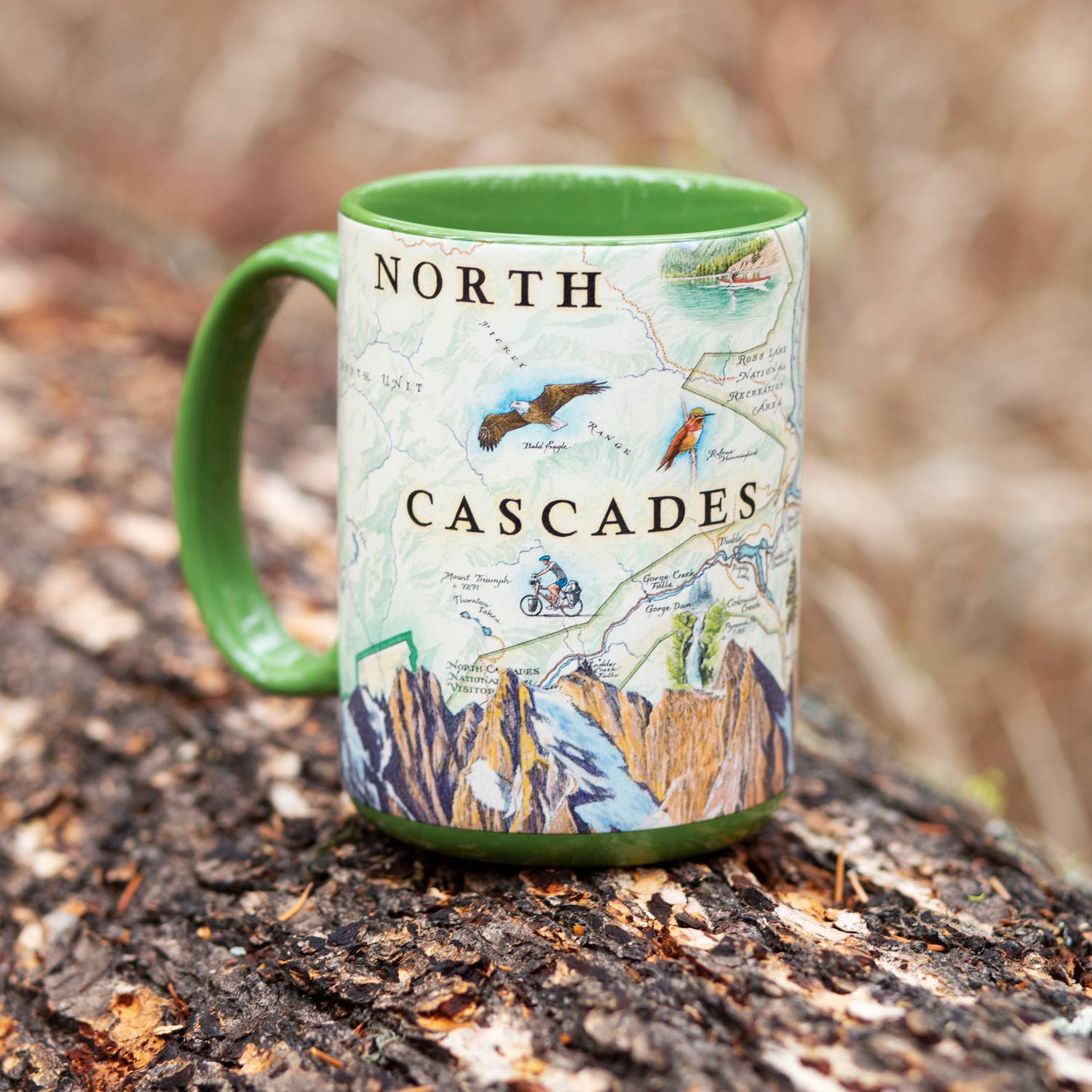 A ceramic mug with a green handle and interior, featuring a detailed map-style illustration labeled 'NORTH CASCADES' with mountains, birds, and outdoor scenes, resting on a tree stump outdoors.