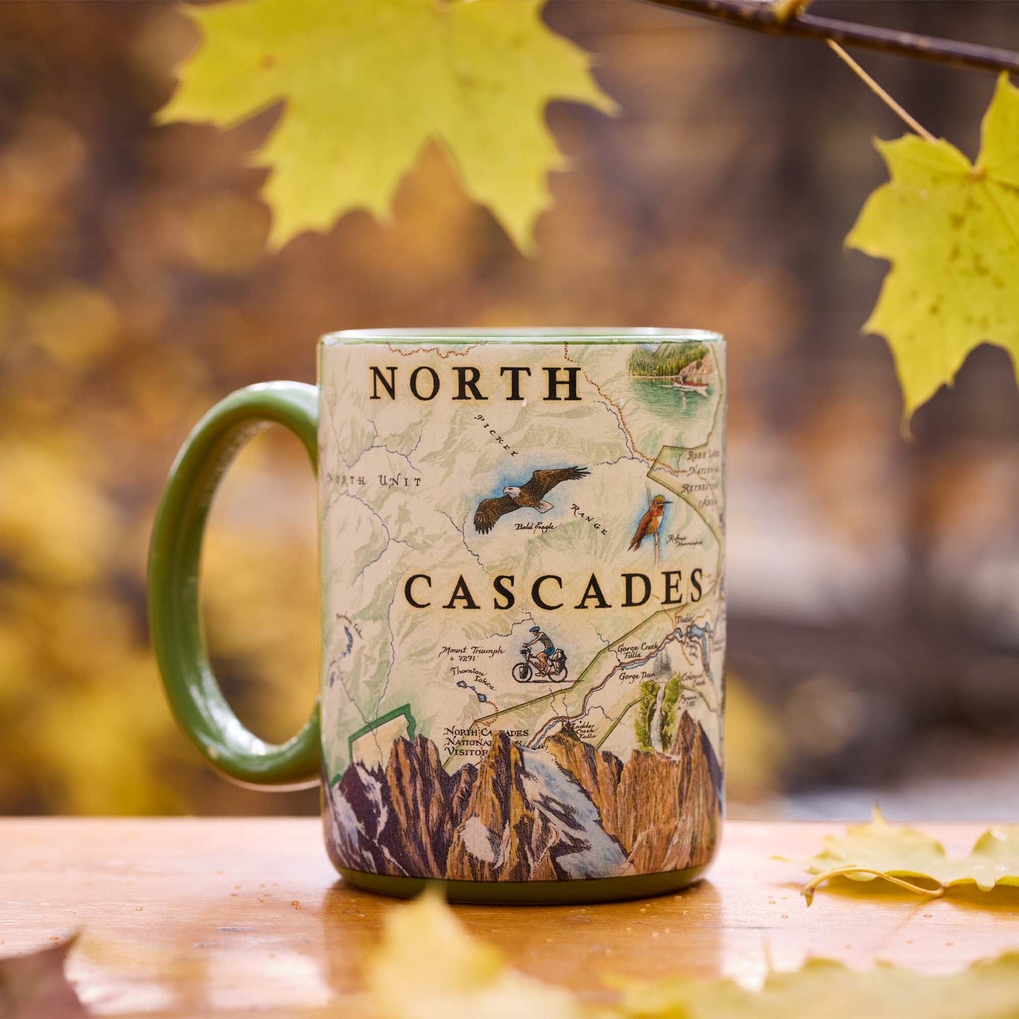 A ceramic mug with a green handle and green rim, printed with a vintage-style map of the North Cascades mountains. The mug is placed on a wooden surface with autumn maple leaves in the background.