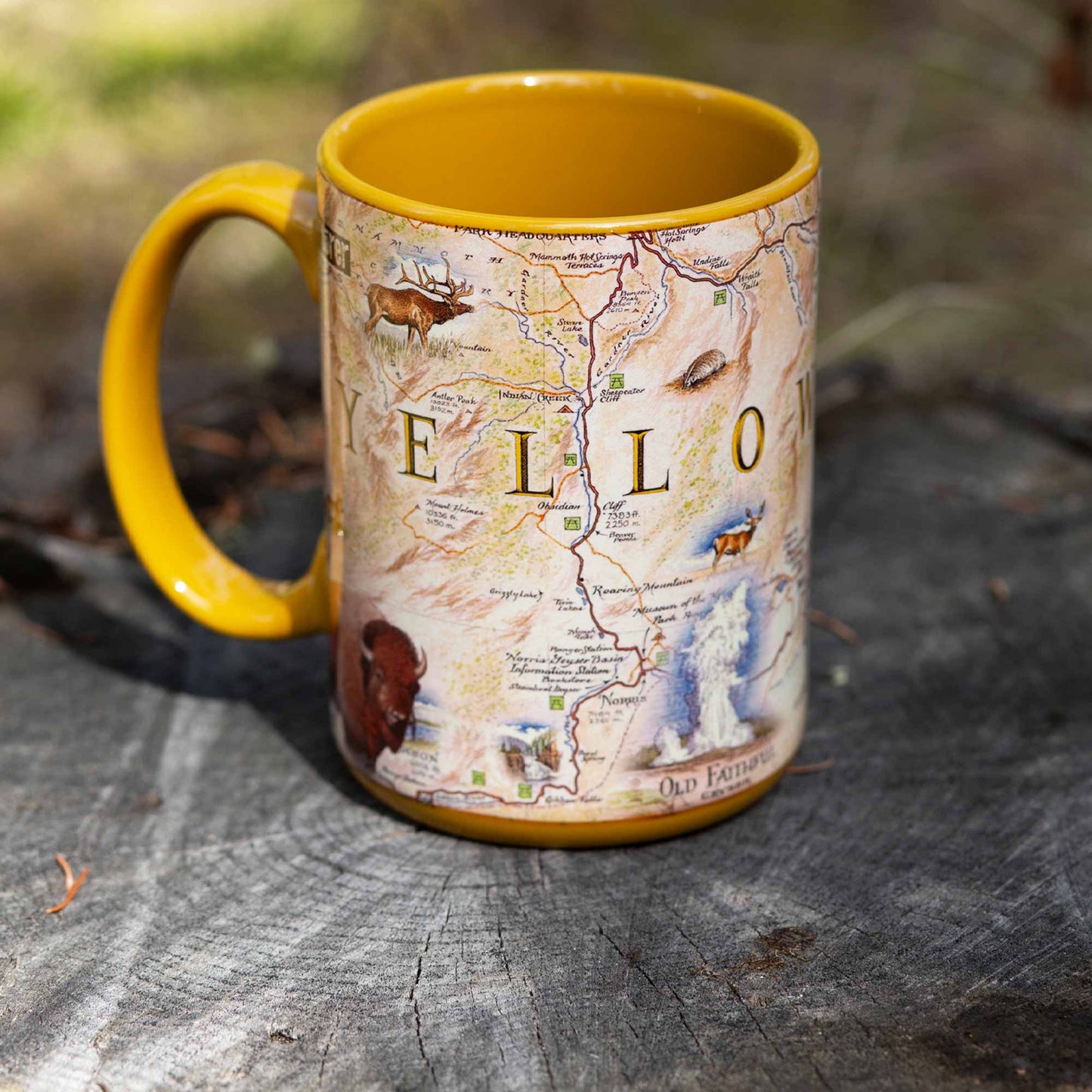 A yellow ceramic mug featuring a vintage-style map of Yellowstone National Park with illustrations of local wildlife, resting on a weathered tree stump outdoors.