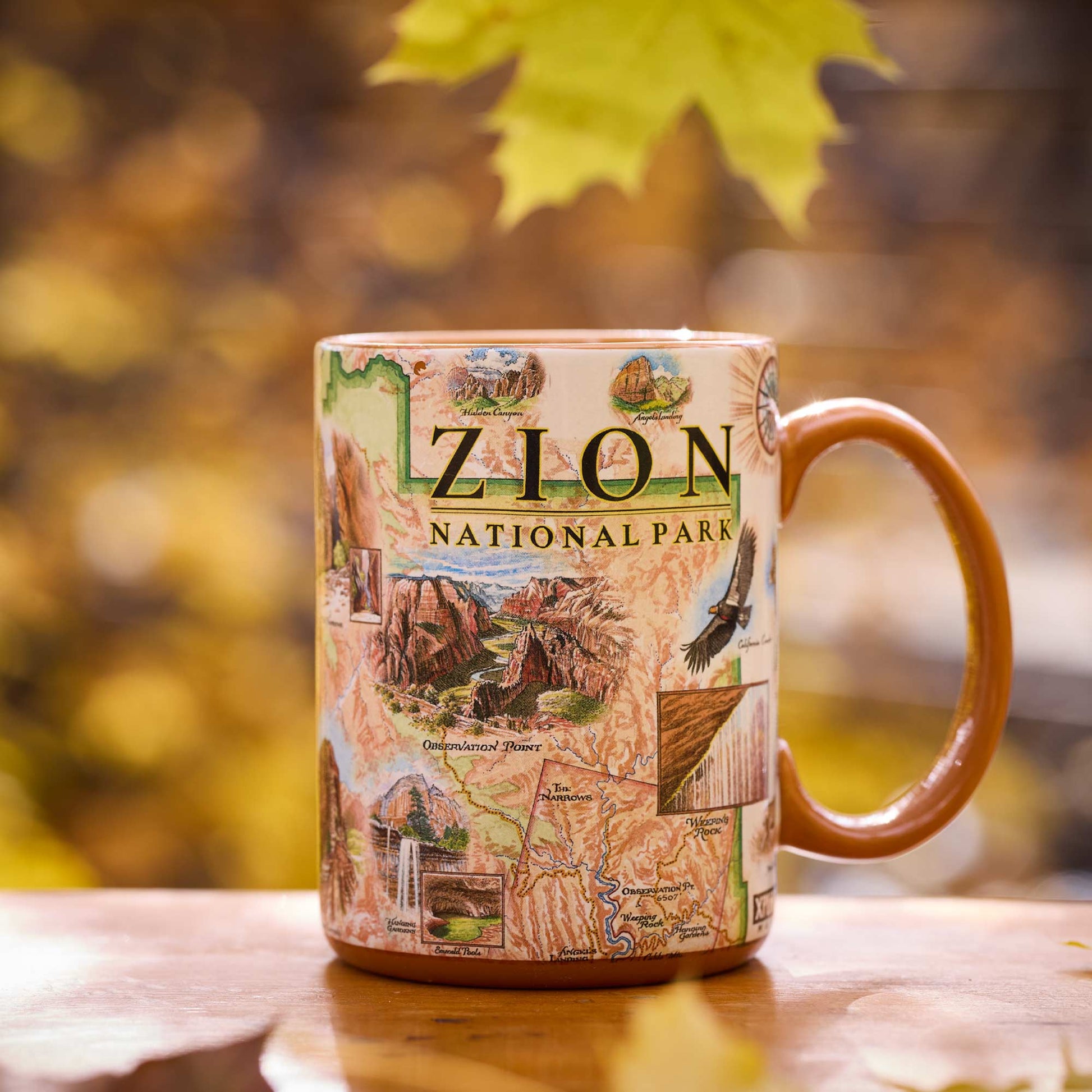 A ceramic mug featuring a detailed map illustration of Zion National Park, set on a wooden surface with a blurred autumn background of yellow leaves.