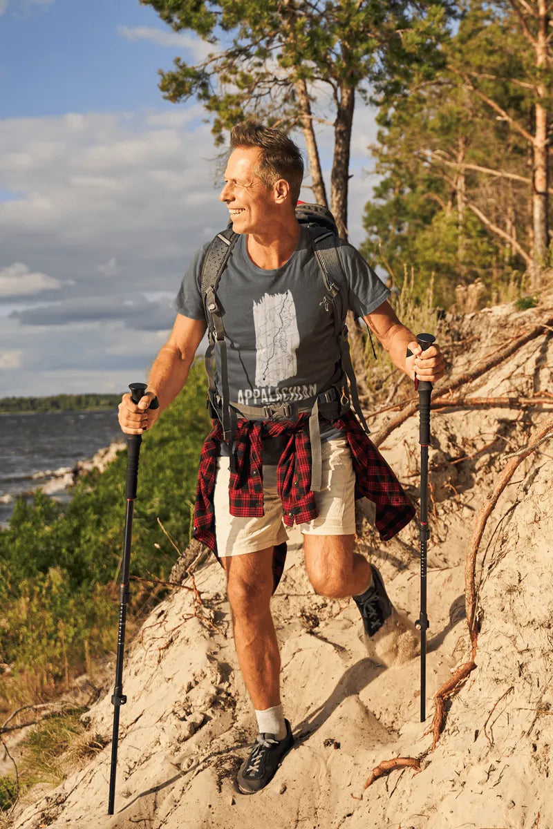 Man hiking on a trail with a backpack and walking sticks, surrounded by trees and water.