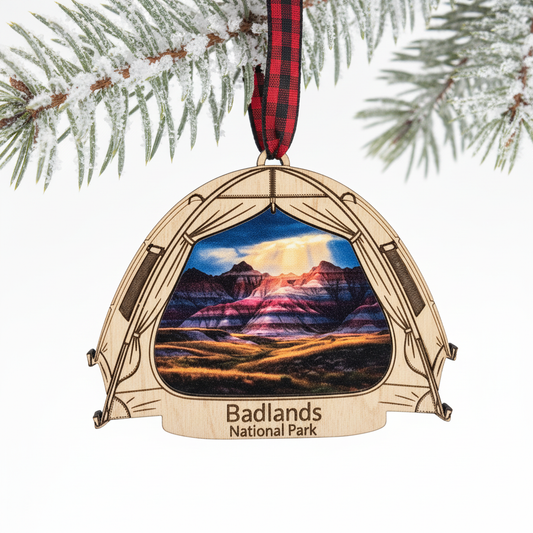 A laser-cut wooden Christmas ornament shaped like a tent, featuring a printed landscape of Badlands National Park and a red plaid hanging ribbon, shown hanging from a snowy pine branch against a white background.