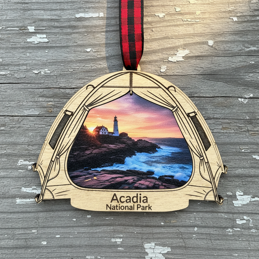 A wooden Acadia National Park ornament shaped like a tent, featuring a printed lighthouse scene with a sunset sky and ocean. The ornament hangs from a red and black plaid ribbon on a weathered gray wood background.