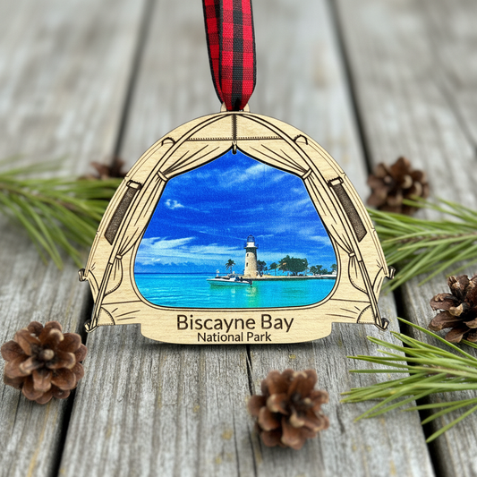 A wooden Christmas ornament shaped like a tent , featuring a printed scenic view of Biscayne Bay National Park with turquoise water, a lighthouse, and a red plaid ribbon hanging from the top. The ornament is displayed on a weathered gray wooden surface with pine needles and pinecones.