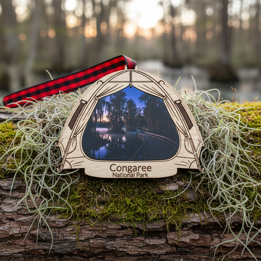 A wooden ornament shaped like a tent, featuring a photo of a forest lake at dusk and the engraved text 'Congaree National Park.' It hangs from a red-and-black buffalo plaid ribbon and is displayed on mossy logs outdoors.