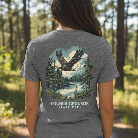 Female walking down a wooded trail  wearing a gray T-shirt with an eagle and forest graphic with the text Council Grounds State Park.