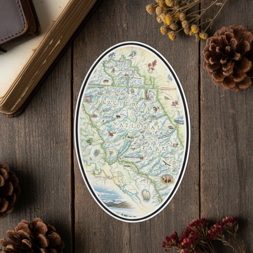 An oval sticker featuring a vintage-style map illustration, displayed on a dark rustic wooden surface surrounded by decorative props including pinecones, dried flowers, an old book, and a leather notebook.