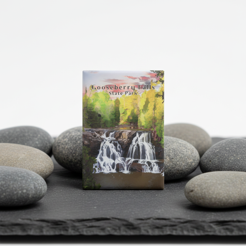 A small rectangular magnet featuring Gooseberry Falls State Park scenery, standing upright on a dark slate surface surrounded by smooth gray stones against a white background.