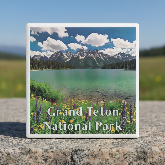 A square coaster featuring a Grand Teton National Park landscape with mountains, a turquoise lake, clouds, and wildflowers, standing on a stone surface outdoors.