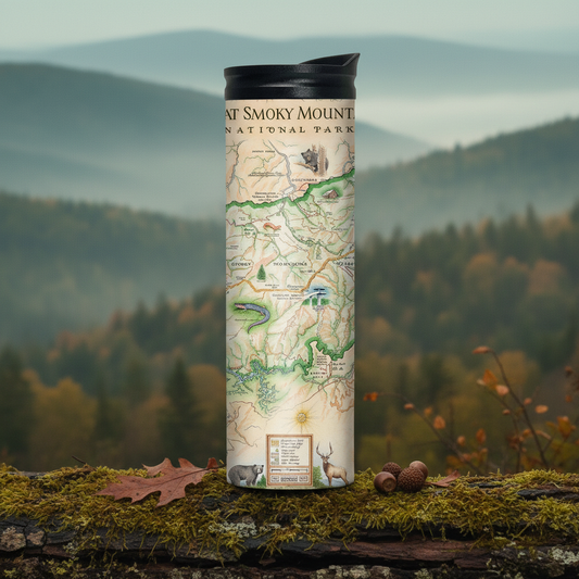A cylindrical insulated tumbler with a black lid stands on a mossy log outdoors, featuring a printed map design labeled Grand Smoky Mountain National Park. The background shows a misty mountain and forest landscape.
