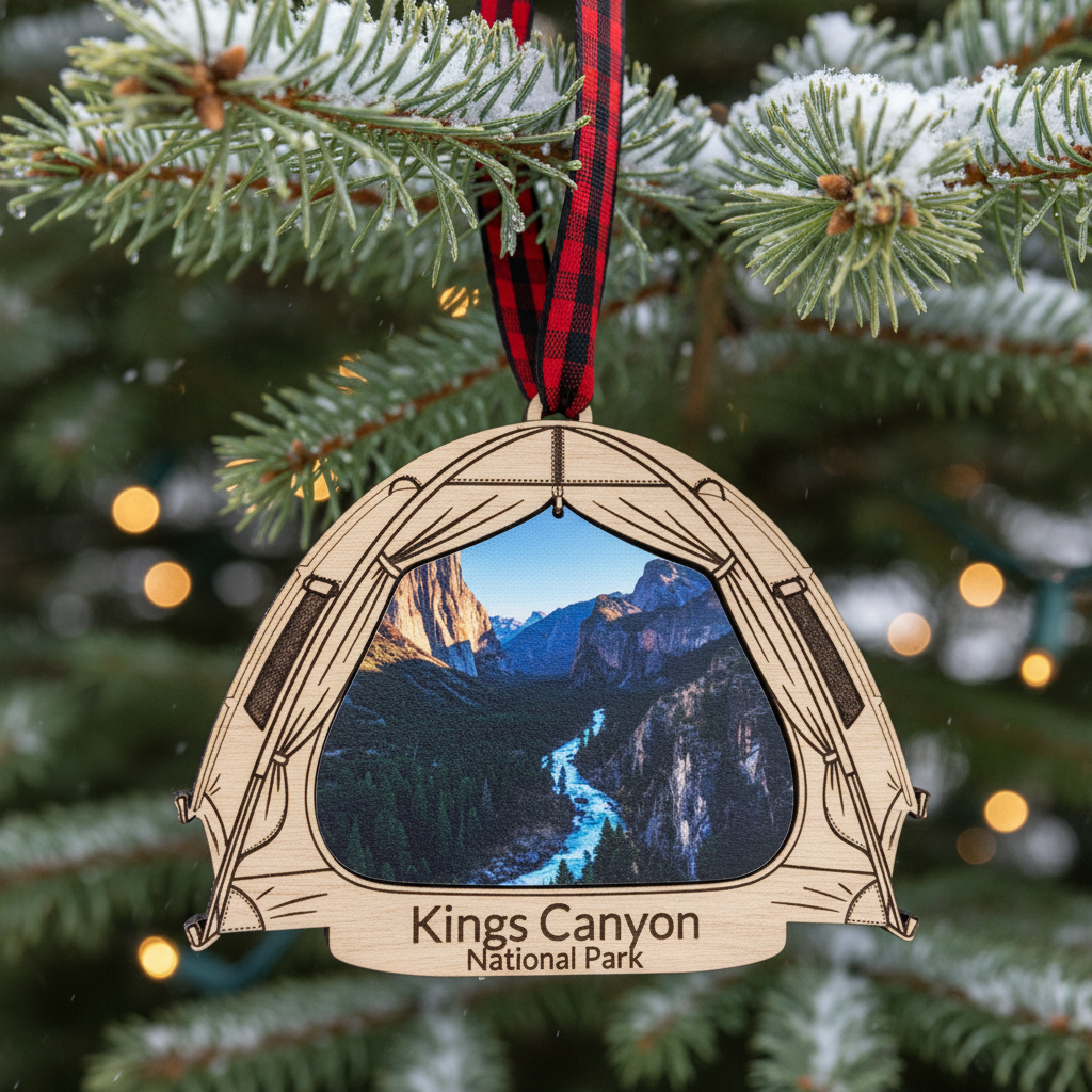 A wooden Christmas ornament shaped like a tent, hanging from a red and black plaid ribbon on a snowy Christmas tree. The ornament features an inset photo of Kings Canyon National Park and the text 'Kings Canyon National Park' engraved at the bottom.