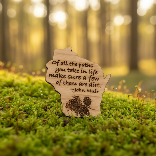A small wooden magnet shaped like the state of Wisconsin stands in green moss outdoors, with a blurred forest background. The wood is engraved with the quote, 'Of all the paths you take in life make sure a few of them are dirt.