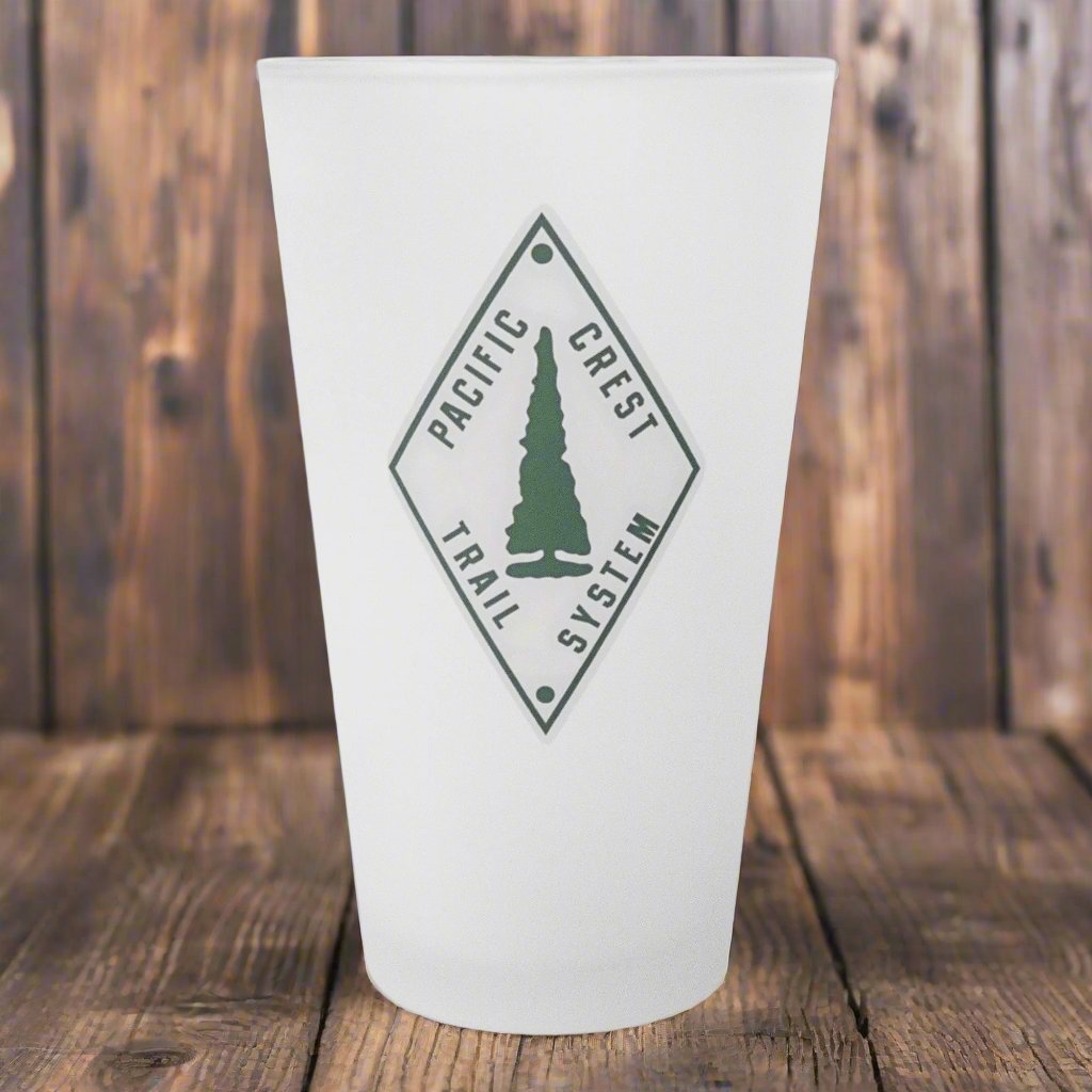 Frosted glass with the Pacific Crest Trail logo sitting on a wood surface. 