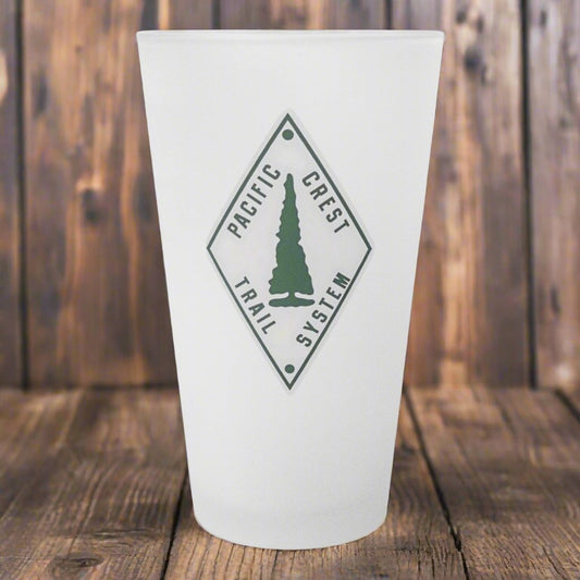Frosted glass with the Pacific Crest Trail logo sitting on a wood surface. 