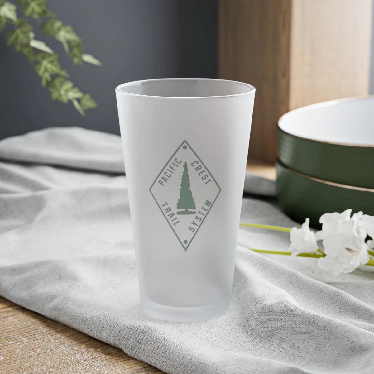 Frosted glass tumbler with the Pacific Crest Trail logo design on a gray cloth surface.
