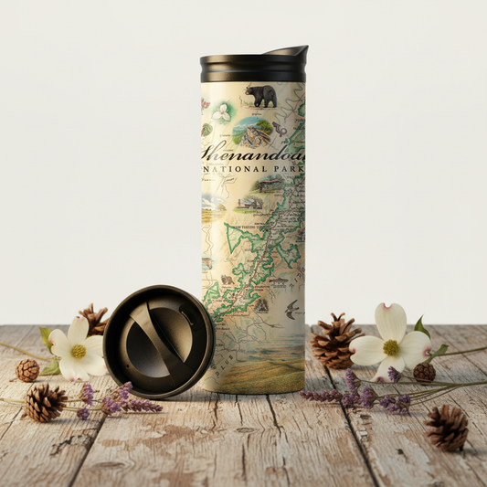 A tall insulated travel tumbler with a lid sits on a rustic wooden surface, with the lid placed beside it. The tumbler features an illustrated map design labeled 'Shenandoah National Park' with vintage-style drawings and text.