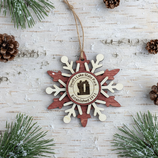 Wooden snowflake ornament with Devils Lake State Park logo on a light wooden obackground with pine branches and cones.