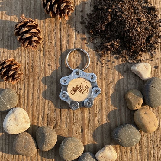 Keychain with bicycle and Bigfoot design on a wooden surface with pine cones, soil, and stones.