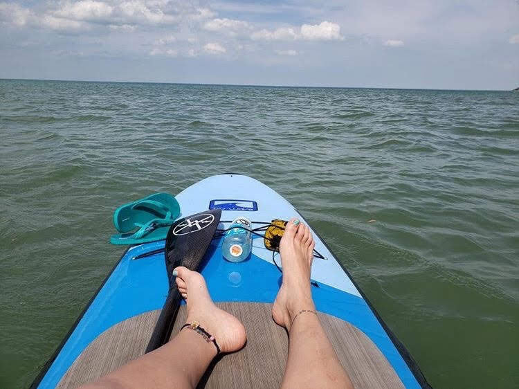 Person paddleboarding on a calm body of water with a clear sky. Displaying anklets